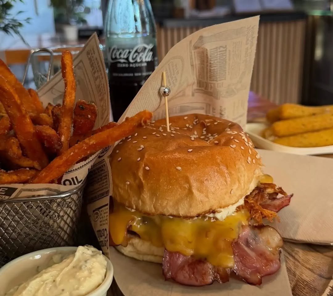 Hambúrguer com queijo derretido e bacon, ao lado de um cesto de batata-doce frita e uma taça de molho